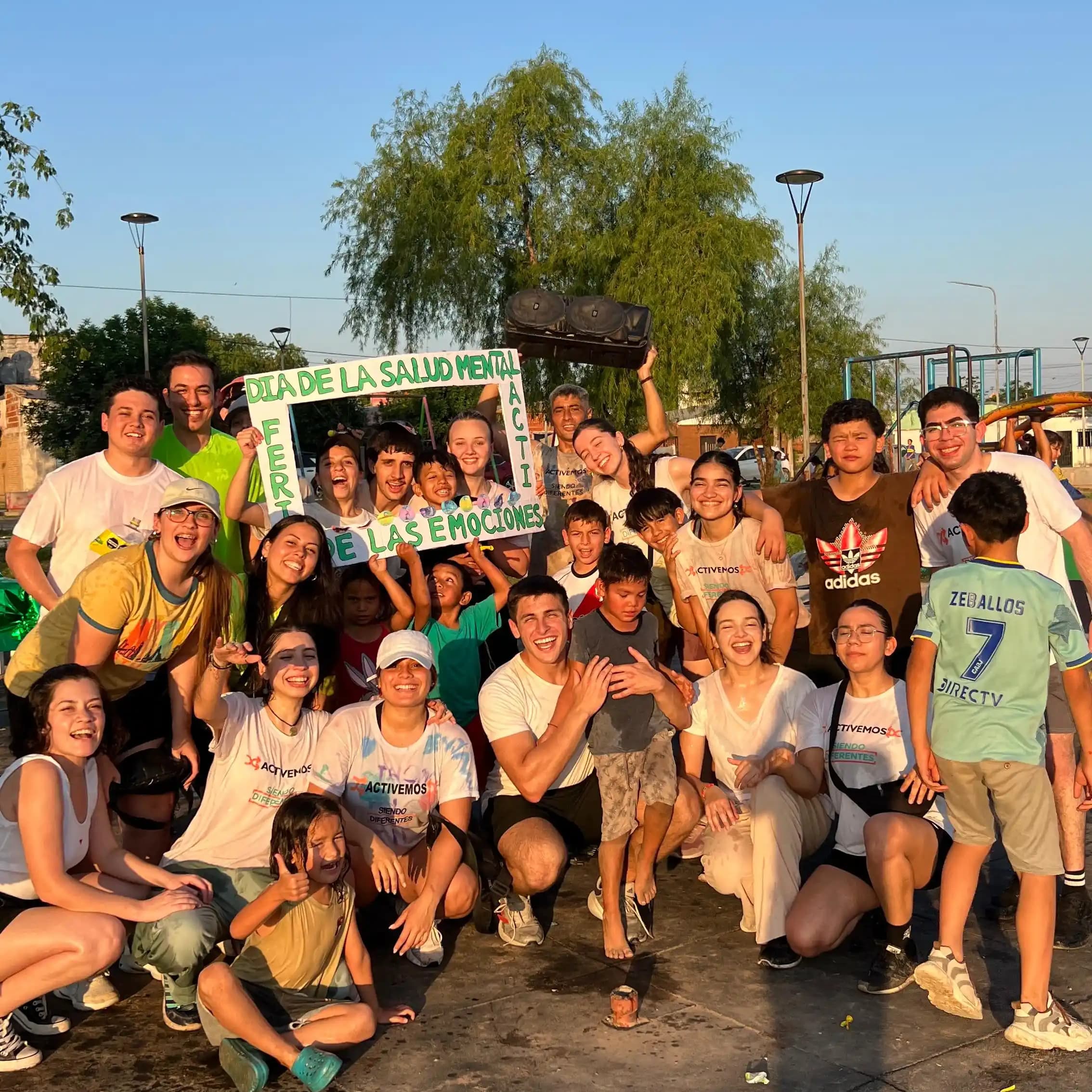 Grupo de chicos, chicas y personas adultas reunidos en una plaza, junto a un marco con letras de colores.